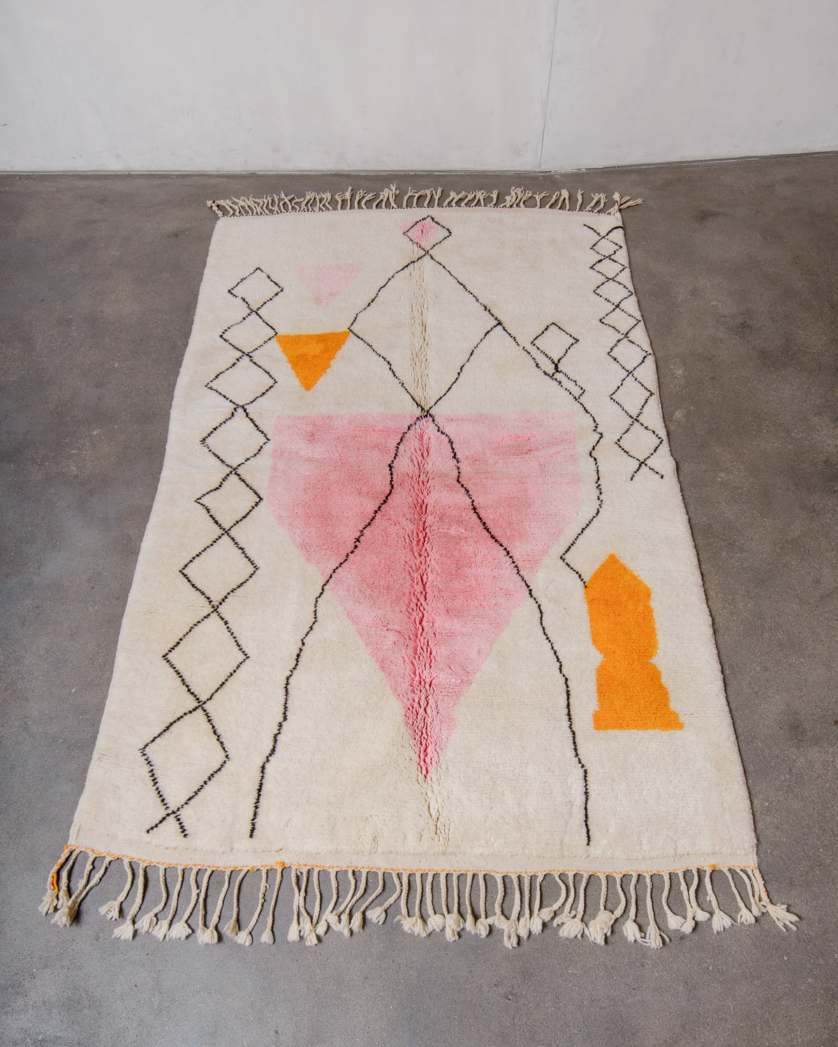 Beniourain Rug made of plush cream wool with a design in pink, orange and black laying flat on concrete floor.
